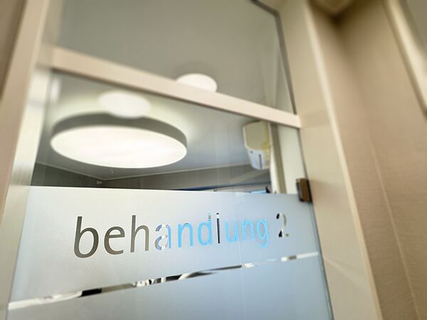 Glastür - das Wort Behandlung ist auf Dekorfollie geplottet und darauf geklebt wordedn - der Blick fällt durch die Tür auf die Lampen an der Praxisdecke im Behandlungsraum 2